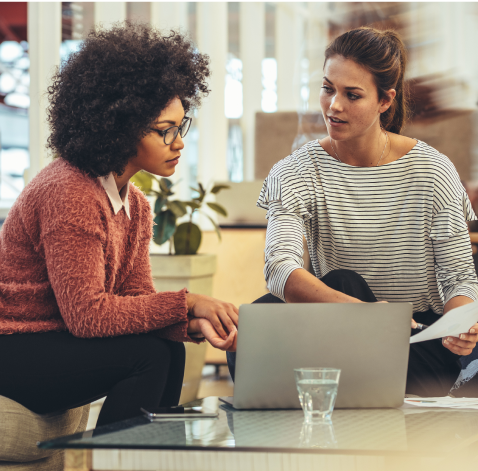 Two women sitting together in an office space, reviewing information on a laptop.
