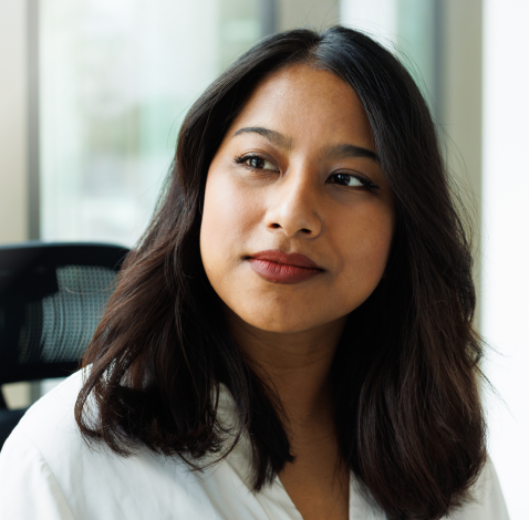 Woman in white shirt, seated indoors with windows and office chair in background.