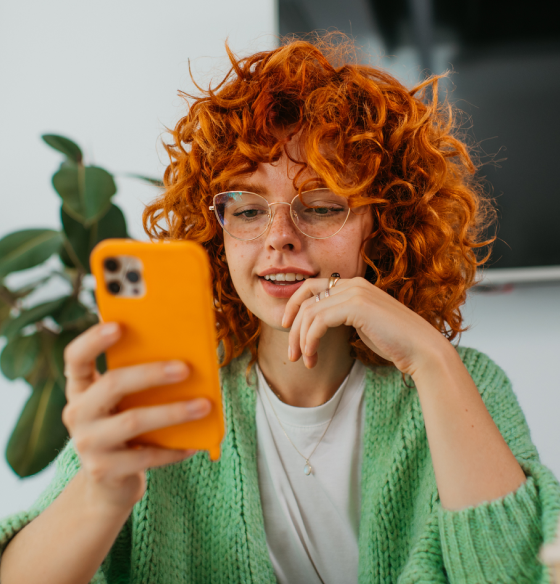 Person with curly red hair and glasses, wearing a green cardigan, holding an orange smartphone; face blurred, plant in background.
