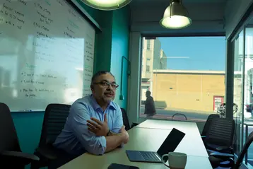 Person sitting at a conference table with a laptop, whiteboard with notes in the background, and a window showing a street view